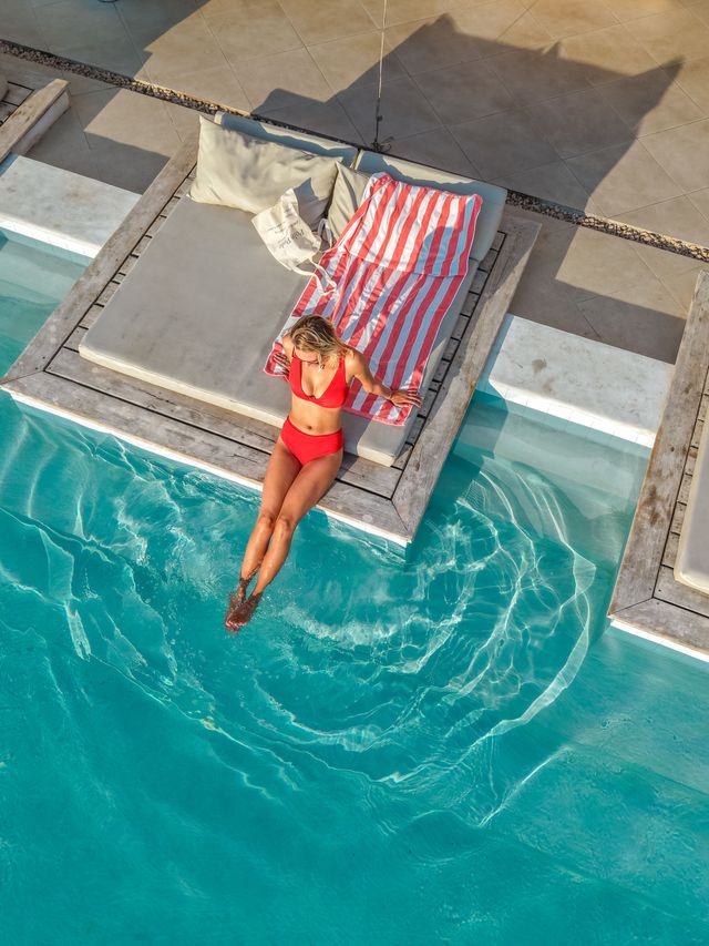 The photo shows a woman relaxing on a comfortable poolside daybed. Wearing a red swimsuit, she dips her feet into the crystal-clear water while enjoying the sunny weather.