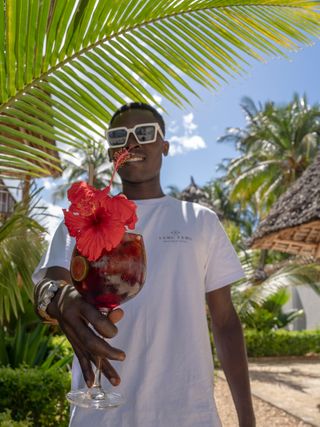 The photo shows a man carrying an elegant tropical cocktail decorated with a red flower. In the background, there are palm trees, exotic greenery, and stylish resort buildings.