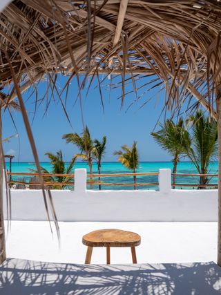 The photo shows a cozy covered relaxation area overlooking the turquoise ocean. The natural thatched roof and light white curtains give the place an exotic and calming character. In the background, palm trees and a blue sky create a peaceful holiday atmosphere.