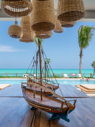 The photo shows an elegant open-air lounge area with a view of a tropical coastline. In the foreground, there is a large wooden table with a decorative model sailboat placed on it. Above the table hang woven pendant lamps in a natural, rustic style. In the background, there is a swimming pool surrounded by sun loungers and palm trees swaying in the wind. Beyond the pool.