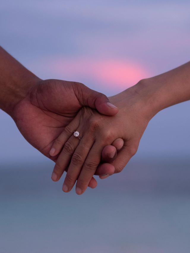 The photo shows two people holding hands against the background of a calm sky with pastel colors. A ring is visible on one of the fingers.