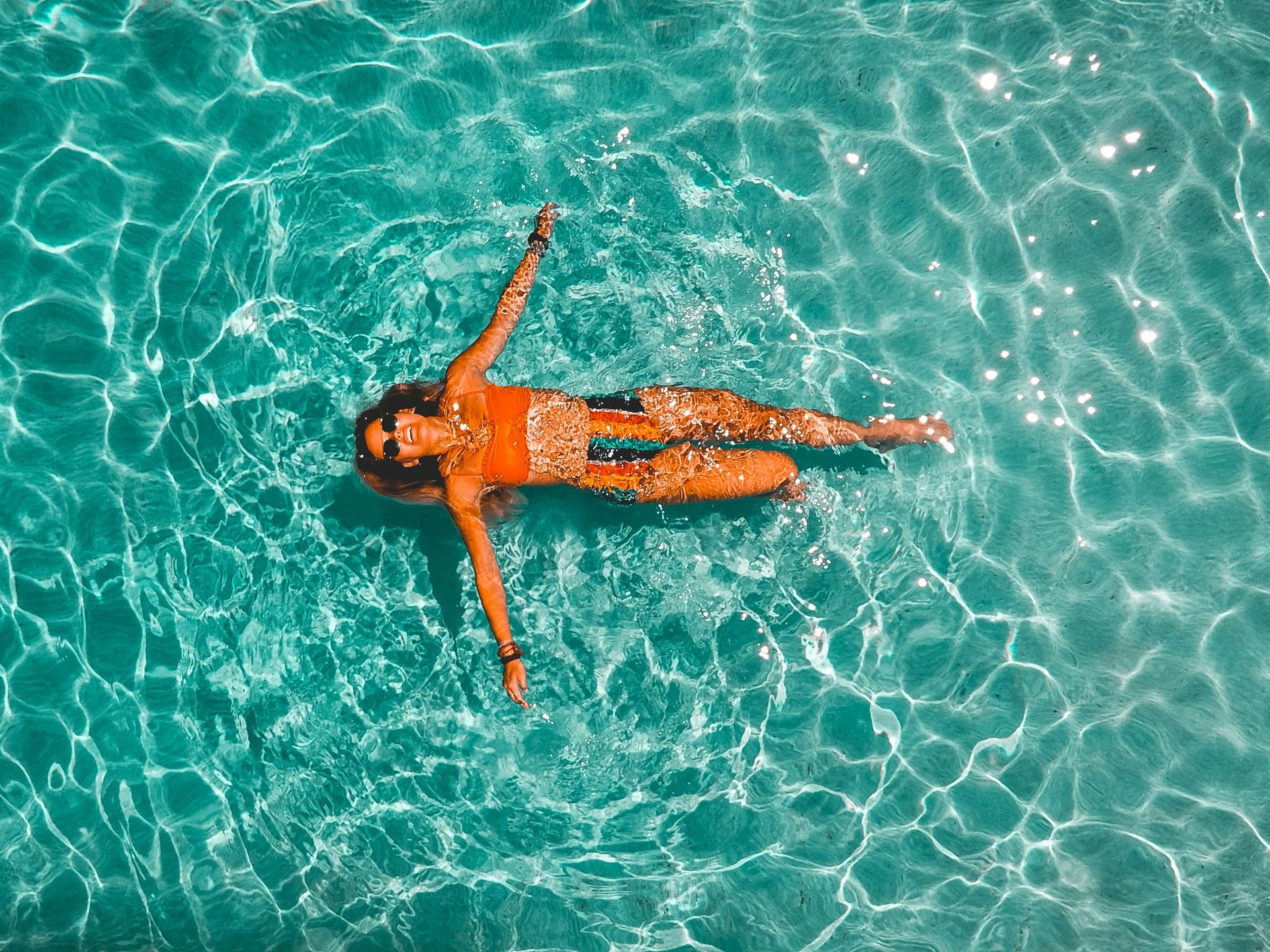 The photo shows a woman floating on her back in crystal-clear turquoise water. Taken from above, it captures the striking light reflections on the water's surface and the gentle waves forming around the swimmer. The sunny weather and vibrant colors highlight the tropical vibe of the place.